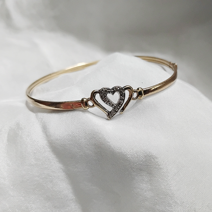 A gold bangle bracelet with a box clasp w/safety latch, featuring two interlocking hearts at the center, one of which is adorned with small, sparkling stones. The bracelet rests on a soft white fabric background, highlighting its delicate and romantic design from Violet Dreamer.