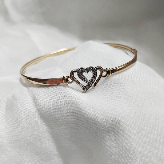 A gold bangle bracelet with a box clasp w/safety latch, featuring two interlocking hearts at the center, one of which is adorned with small, sparkling stones. The bracelet rests on a soft white fabric background, highlighting its delicate and romantic design from Violet Dreamer.