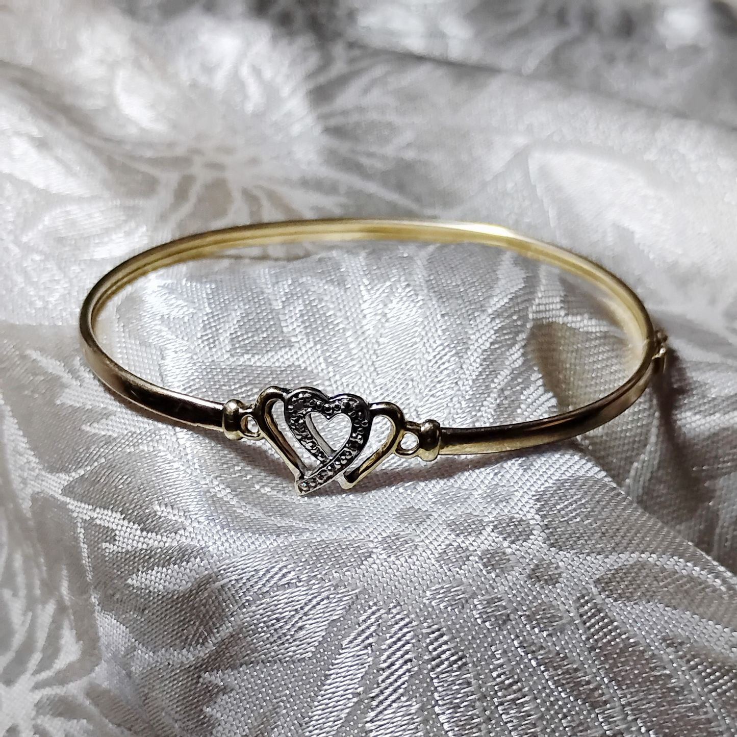 A gold bangle bracelet with a box clasp w/safety latch, featuring two interlocking hearts at the center, one of which is adorned with small, sparkling stones. The bracelet rests on a soft white fabric background, highlighting its delicate and romantic design from Violet Dreamer.