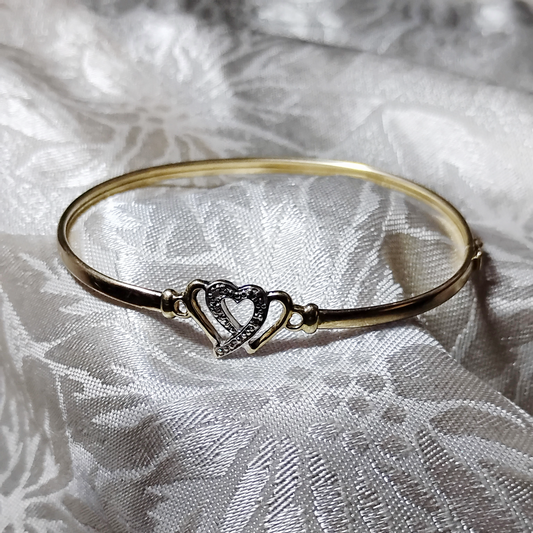 A gold bangle bracelet with a box clasp w/safety latch, featuring two interlocking hearts at the center, one of which is adorned with small, sparkling stones. The bracelet rests on a soft white fabric background, highlighting its delicate and romantic design from Violet Dreamer.