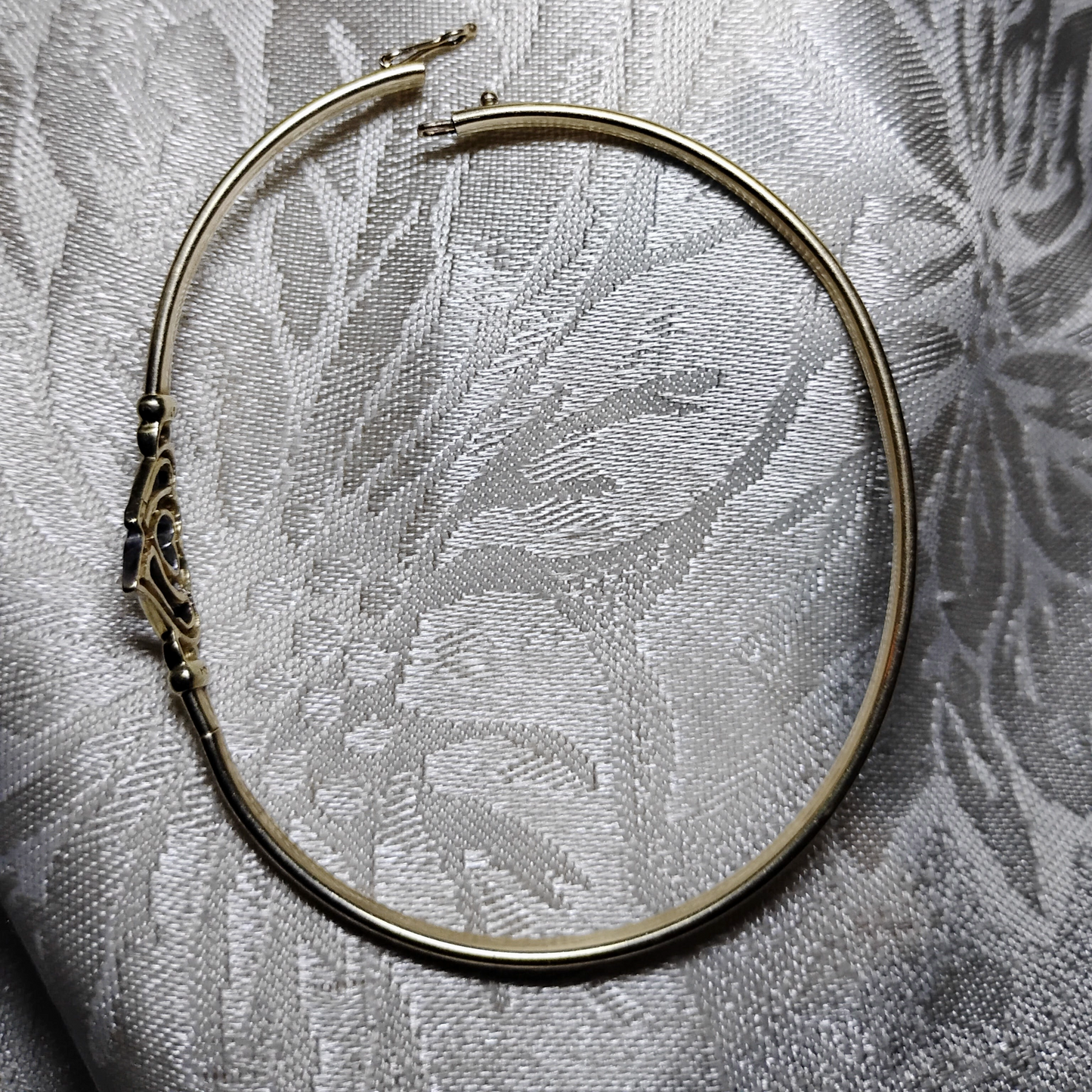 An open, gold bangle bracelet with a box clasp w/safety latch, featuring two interlocking hearts at the center, one of which is adorned with small, sparkling stones. The bracelet rests on a soft white fabric background, highlighting its delicate and romantic design from Violet Dreamer.