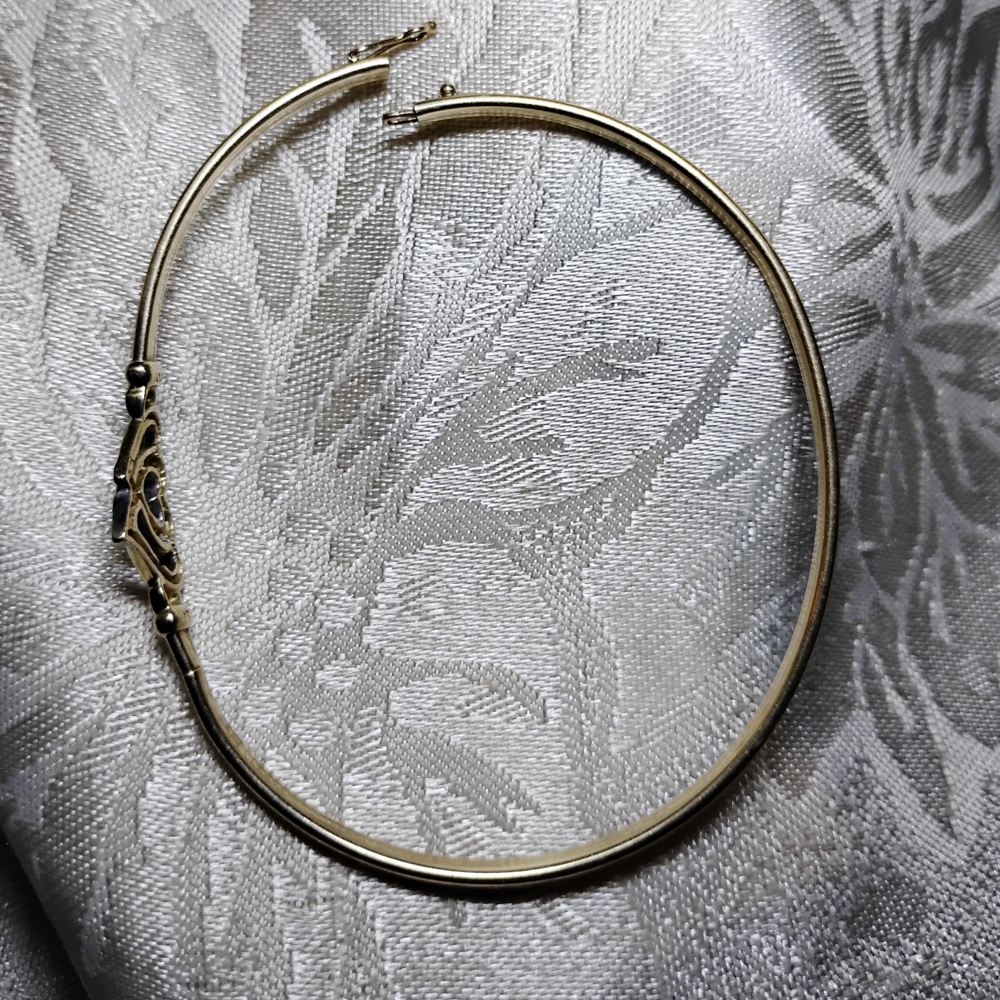 An open, gold bangle bracelet with a box clasp w/safety latch, featuring two interlocking hearts at the center, one of which is adorned with small, sparkling stones. The bracelet rests on a soft white fabric background, highlighting its delicate and romantic design from Violet Dreamer.