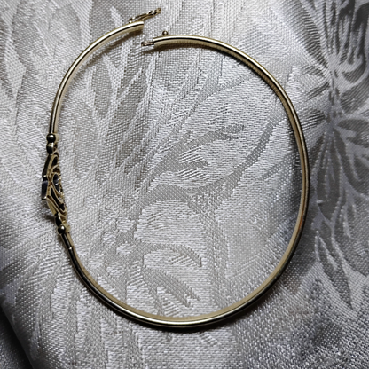 An open, gold bangle bracelet with a box clasp w/safety latch, featuring two interlocking hearts at the center, one of which is adorned with small, sparkling stones. The bracelet rests on a soft white fabric background, highlighting its delicate and romantic design from Violet Dreamer.