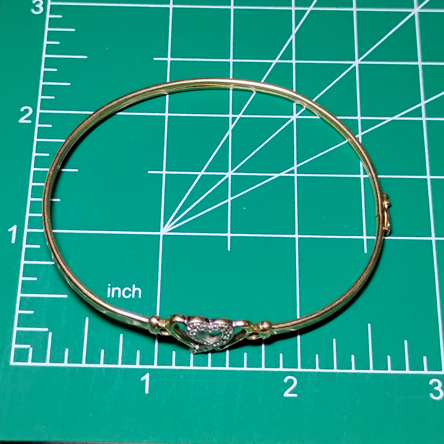 A gold bangle bracelet with a box clasp w/safety latch, featuring two interlocking hearts at the center, one of which is adorned with small, sparkling stones. Bracelet is on a measuring mat showing diameter of approx 2.5 inches.