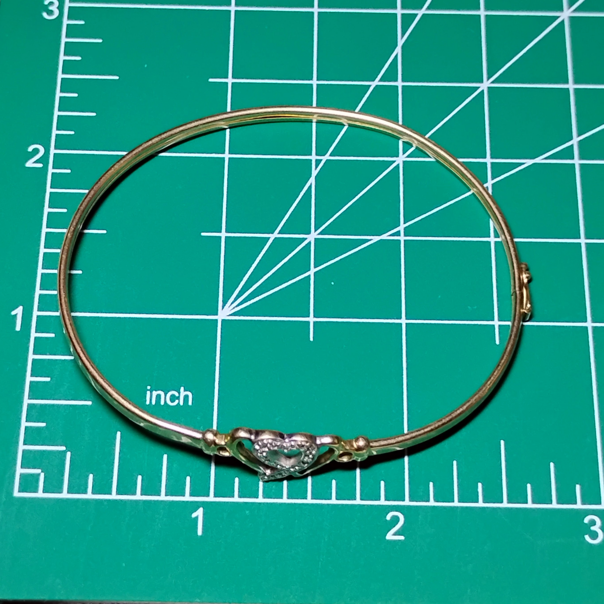 A gold bangle bracelet with a box clasp w/safety latch, featuring two interlocking hearts at the center, one of which is adorned with small, sparkling stones. Bracelet is on a measuring mat showing diameter of approx 2.5 inches.
