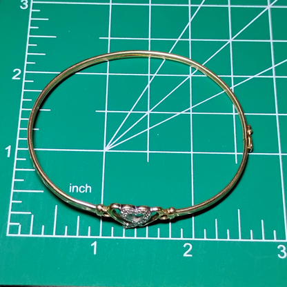 A gold bangle bracelet with a box clasp w/safety latch, featuring two interlocking hearts at the center, one of which is adorned with small, sparkling stones. Bracelet is on a measuring mat showing diameter of approx 2.5 inches.