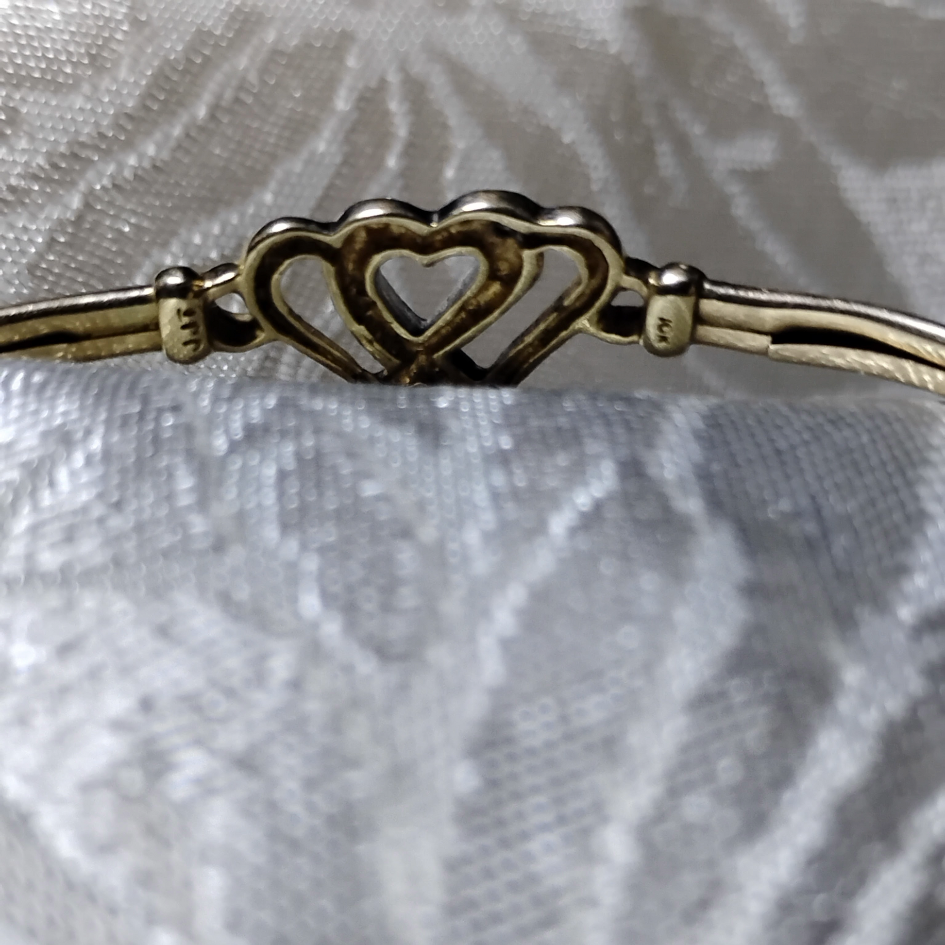Close up view of the back of a gold bangle bracelet with a box clasp w/safety latch, featuring two interlocking hearts at the center, one of which is adorned with small, sparkling stones. The marks "10k" and "JJT" can be seen, from Violet Dreamer.