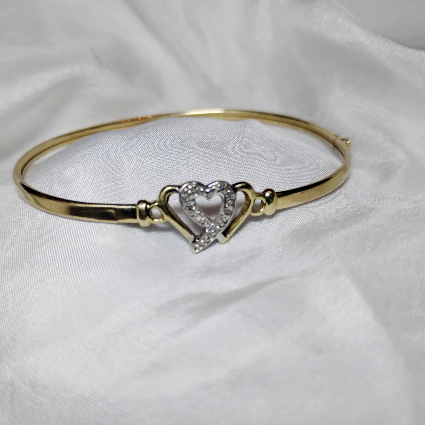 A gold bangle bracelet with a box clasp w/safety latch, featuring two interlocking hearts at the center, one of which is adorned with small, sparkling stones. The bracelet rests on a soft white fabric background, highlighting its delicate and romantic design from Violet Dreamer.