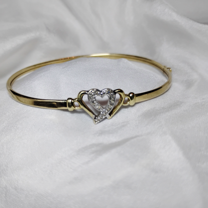 A gold bangle bracelet with a box clasp w/safety latch, featuring two interlocking hearts at the center, one of which is adorned with small, sparkling stones. The bracelet rests on a soft white fabric background, highlighting its delicate and romantic design from Violet Dreamer.