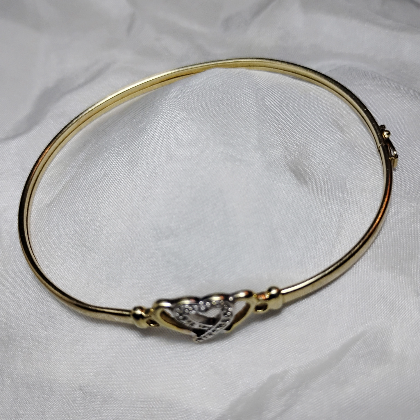A gold bangle bracelet with a box clasp w/safety latch, featuring two interlocking hearts at the center, one of which is adorned with small, sparkling stones. The bracelet rests on a soft white fabric background, highlighting its delicate and romantic design from Violet Dreamer.