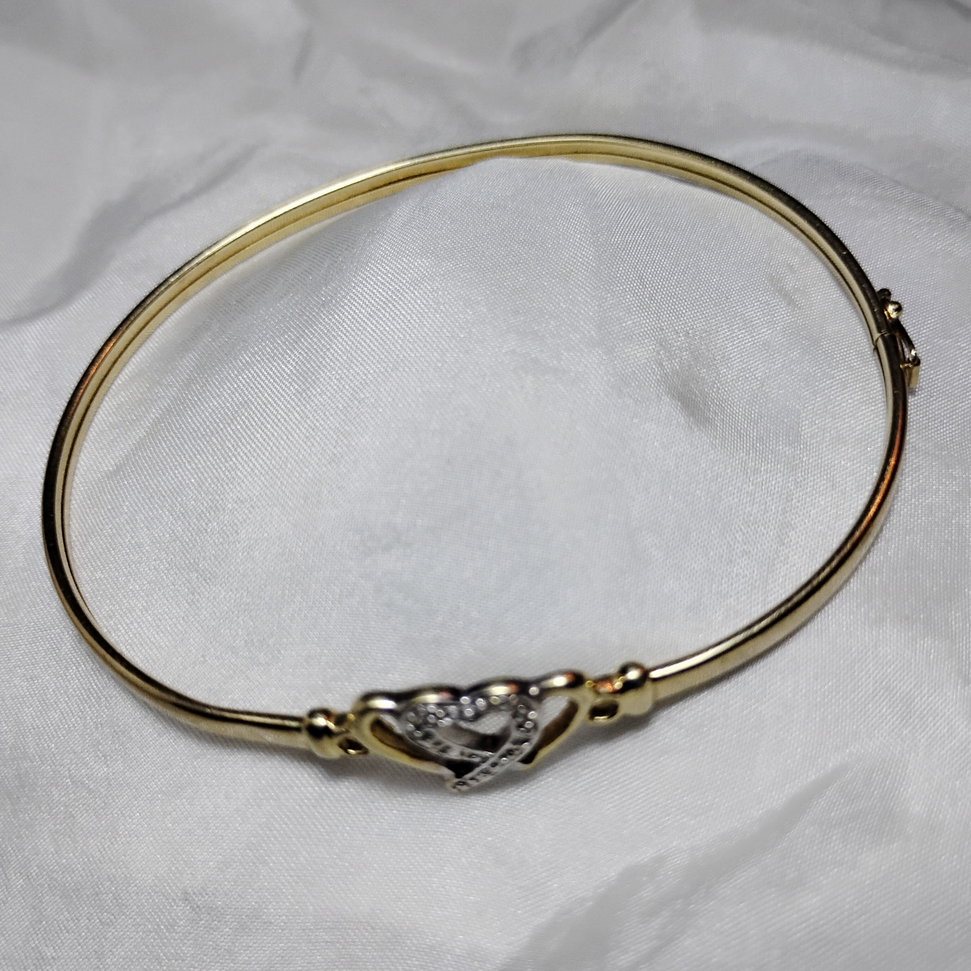 A gold bangle bracelet with a box clasp w/safety latch, featuring two interlocking hearts at the center, one of which is adorned with small, sparkling stones. The bracelet rests on a soft white fabric background, highlighting its delicate and romantic design from Violet Dreamer.