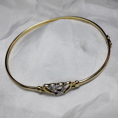 A gold bangle bracelet with a box clasp w/safety latch, featuring two interlocking hearts at the center, one of which is adorned with small, sparkling stones. The bracelet rests on a soft white fabric background, highlighting its delicate and romantic design from Violet Dreamer.