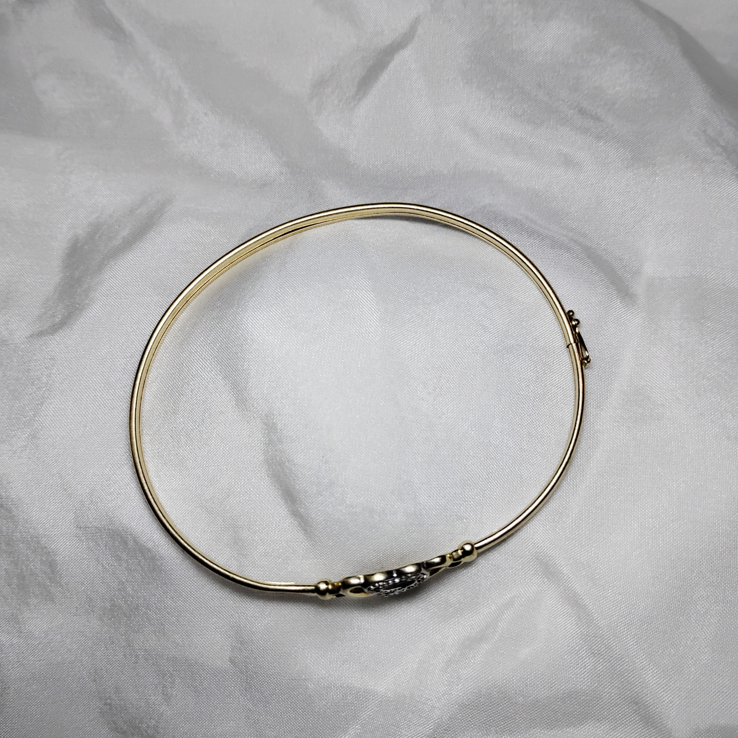 A gold bangle bracelet with a box clasp w/safety latch, featuring two interlocking hearts at the center, one of which is adorned with small, sparkling stones. The bracelet rests on a soft white fabric background, highlighting its delicate and romantic design from Violet Dreamer.