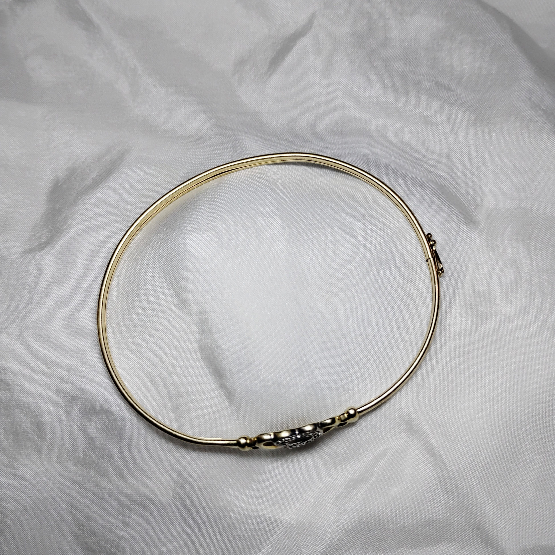 A gold bangle bracelet with a box clasp w/safety latch, featuring two interlocking hearts at the center, one of which is adorned with small, sparkling stones. The bracelet rests on a soft white fabric background, highlighting its delicate and romantic design from Violet Dreamer.