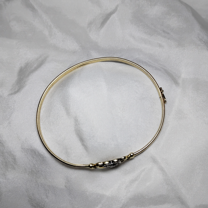 A gold bangle bracelet with a box clasp w/safety latch, featuring two interlocking hearts at the center, one of which is adorned with small, sparkling stones. The bracelet rests on a soft white fabric background, highlighting its delicate and romantic design from Violet Dreamer.