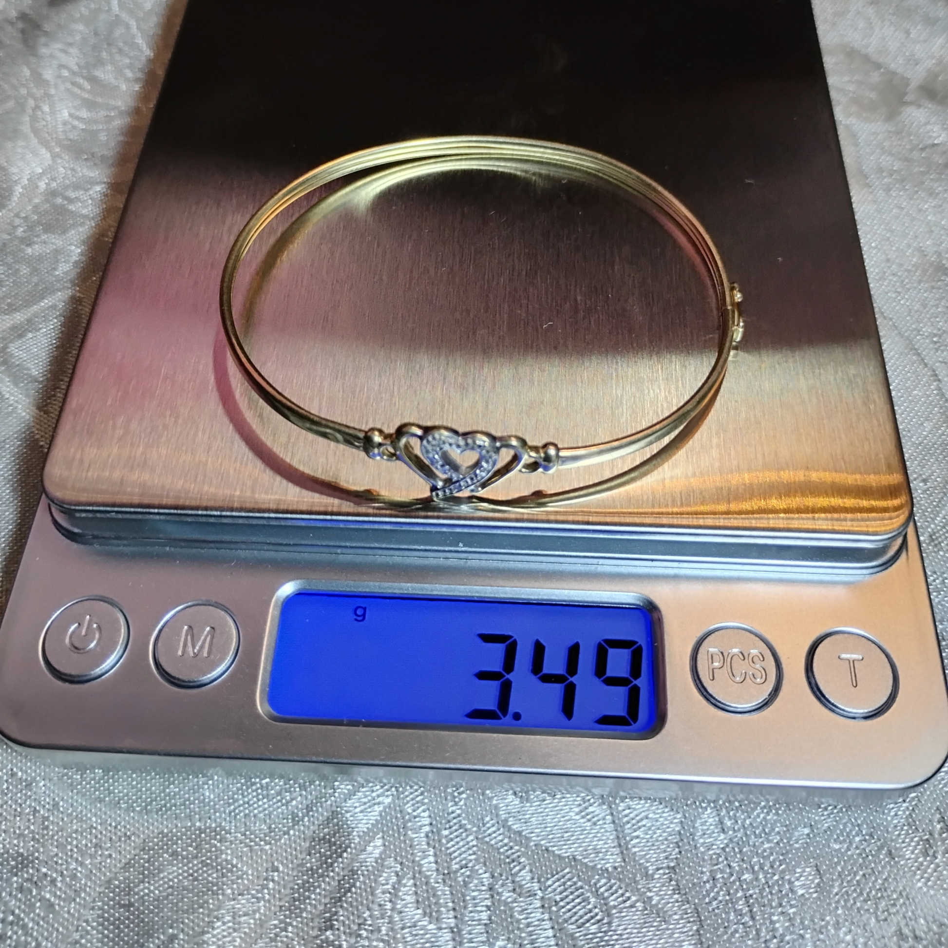 A gold bangle bracelet with a box clasp w/safety latch, featuring two interlocking hearts at the center, one of which is adorned with small, sparkling stones. The bracelet sits on a digital scale with a reading of 3.49g, from Violet Dreamer.
