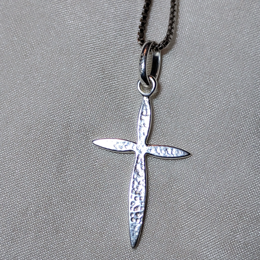 Close-up image of the back of a  polished silver cross pendant with smooth, tapered edges, hanging from a loop on a silver chain, displayed against a light textured fabric background from Violet Dreamer.